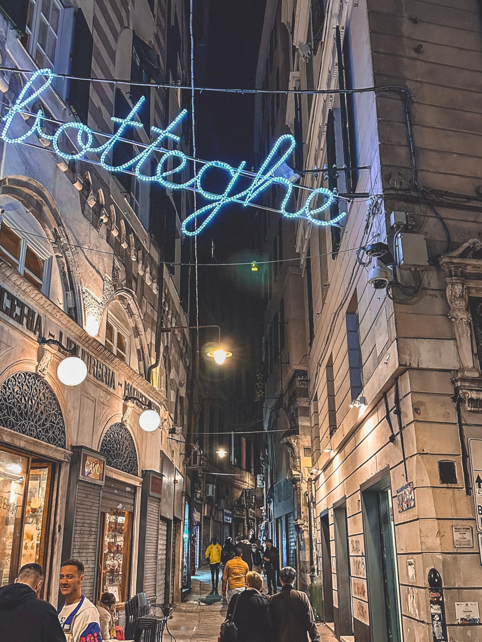 caruggio genovese di sera con persone che passeggiano e una scritta in primo piano illuminata con scritto "botteghe"